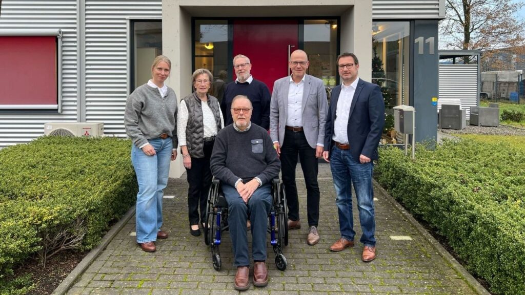 Gruppe von sechs Personen vor einem modernen Gebäude, darunter ein Mann im Rollstuhl und mehrere Erwachsene in formeller Kleidung.