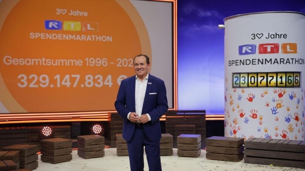 Mann in Anzug steht vor einer Anzeige mit der Gesamtsumme des RTL Spendenmarathons von 1996 bis 2020.