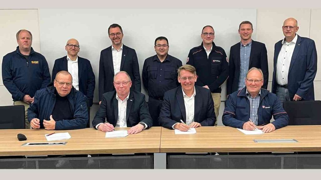 Gruppenfoto von Personen, die an einem Tisch sitzen und Verträge unterzeichnen, im Hintergrund weitere Anwesende.