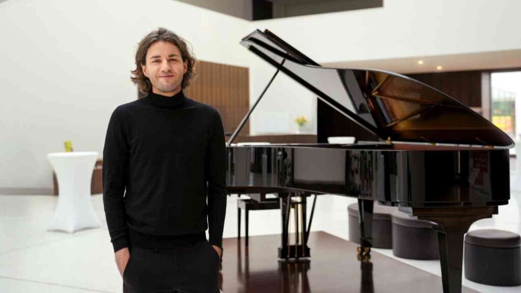 Mann in schwarzem Rollkragenpullover steht neben einem schwarzen Flügelpiano in einem modernen Innenraum.
