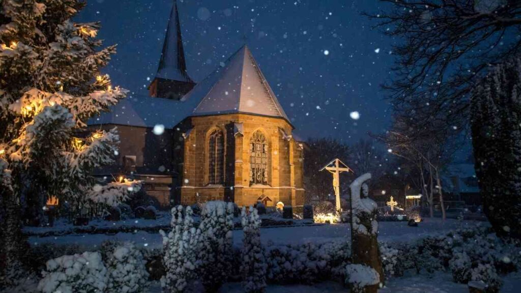St. Vitus Kirche in Lünne, beleuchtet im Schnee bei Nacht mit schneebedecktem Gelände und Weihnachtsdekoration.