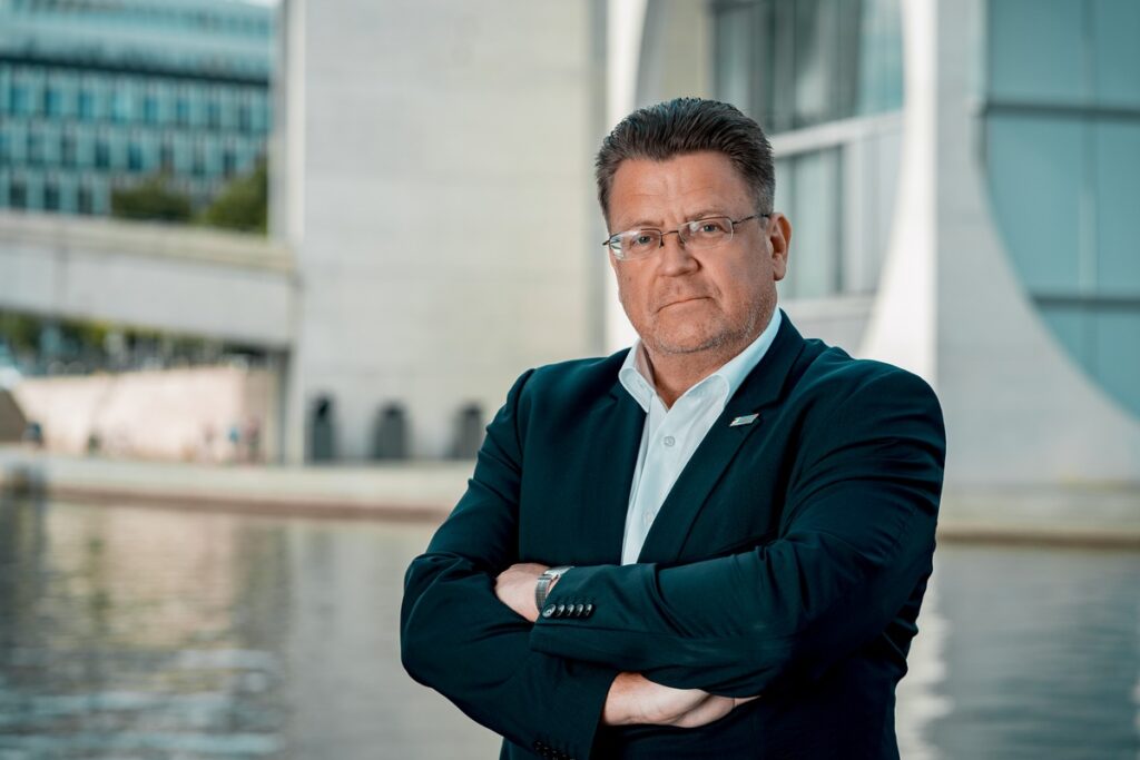 Mann mit Brille und verschränkten Armen in einem dunklen Anzug vor moderner Architektur am Wasser.