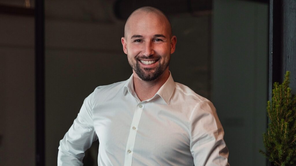 Mann mit kurzem Bart und Glatze, der in einem weißen Hemd lächelt und eine entspannte Pose einnimmt.