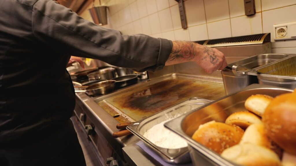 Koch in einer Küche, der mit einer Hand eine Pfanne auf einer heißen Kochplatte anhebt.