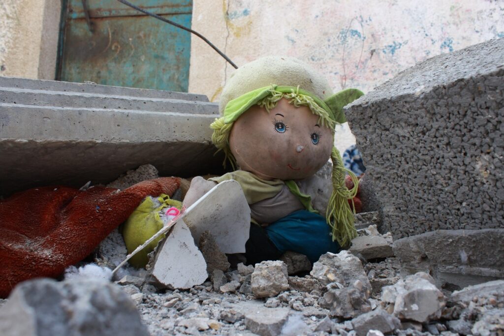 Eine beschädigte Puppe mit grüner Mütze und blonden Haaren, umgeben von Trümmern und Schutt.