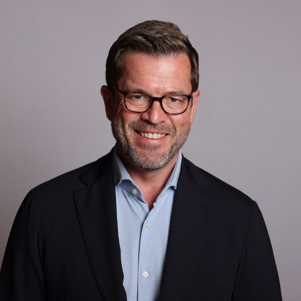 Mann mit Brille und Bart, der in einem dunklen Blazer und einem hellen Hemd lächelt.