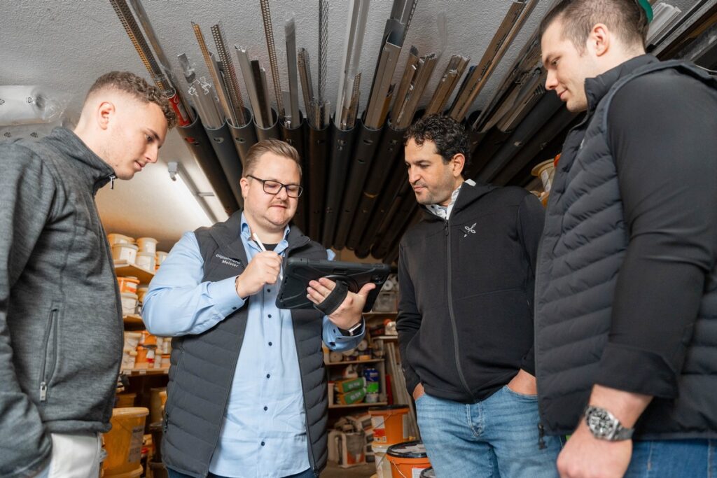 Vier Männer in einem Lagerraum, einer nutzt ein Tablet, während die anderen zuschauen und diskutieren.