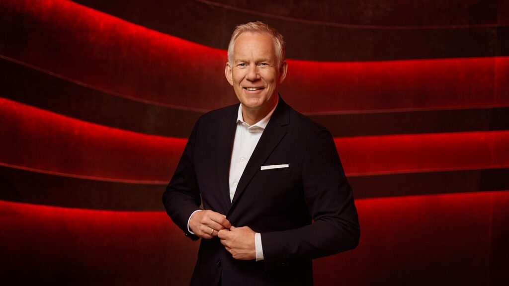 Mann in einem schwarzen Anzug mit weißem Hemd, der an einem roten Hintergrund lächelt und seine Jacke zuknöpft.