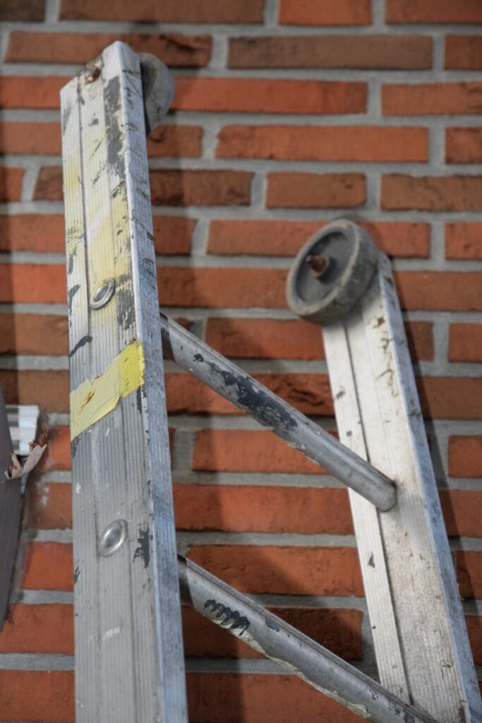 Aluminiumleiter mit abgenutzten Stufen und gelben Markierungen, angelehnt an eine Ziegelwand.