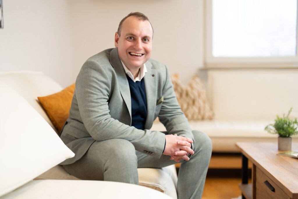 Mann in einem grauen Anzug sitzt auf einem Sofa und lächelt freundlich in die Kamera.