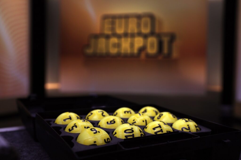 Gelbe Eurojackpot-Lotteriekugeln mit Zahlen in einem schwarzen Behälter vor einem Eurojackpot-Hintergrund.