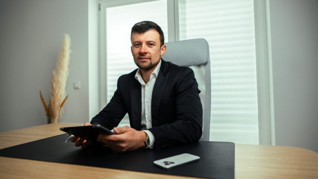 Mann in Anzug sitzt an einem Schreibtisch und hält ein Tablet in der Hand, mit einem Smartphone auf dem Tisch.