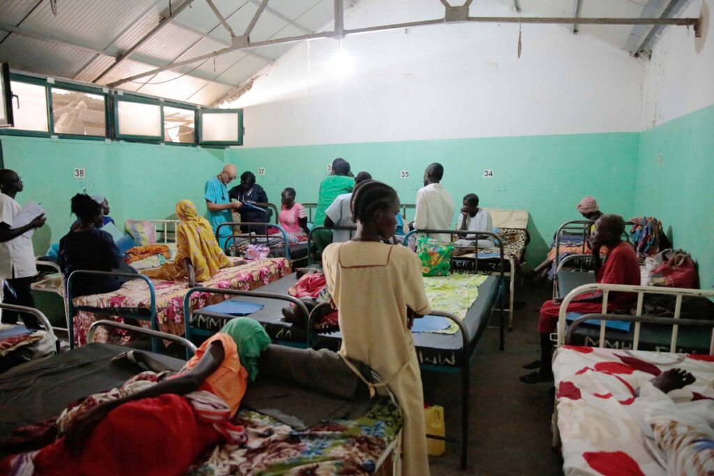 Patienten in einem Krankenhauszimmer mit mehreren Betten und medizinischem Personal, das sich um die Patienten kümmert.