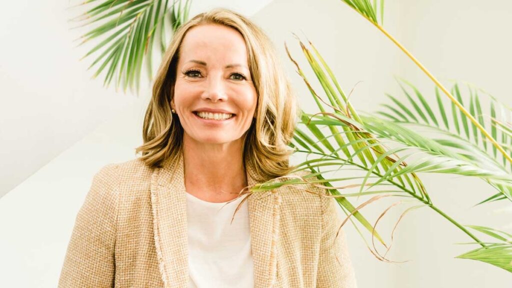 Frau mit blonden Haaren, die in einem hellen Outfit vor grünen Pflanzen steht und lächelt.