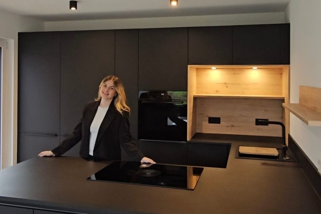 Moderne Küche mit schwarzem Design und Holzregal, präsentiert von einer Frau in einem schwarzen Blazer.