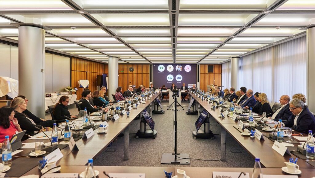 Konferenzraum mit einem langen Tisch, umgeben von Teilnehmern und einem Bildschirm im Hintergrund.