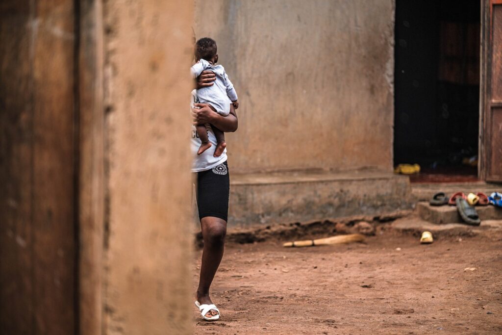 Eine Person hält ein Kleinkind in einem informellen Außenbereich mit erdigen Boden und einfachen Wänden.