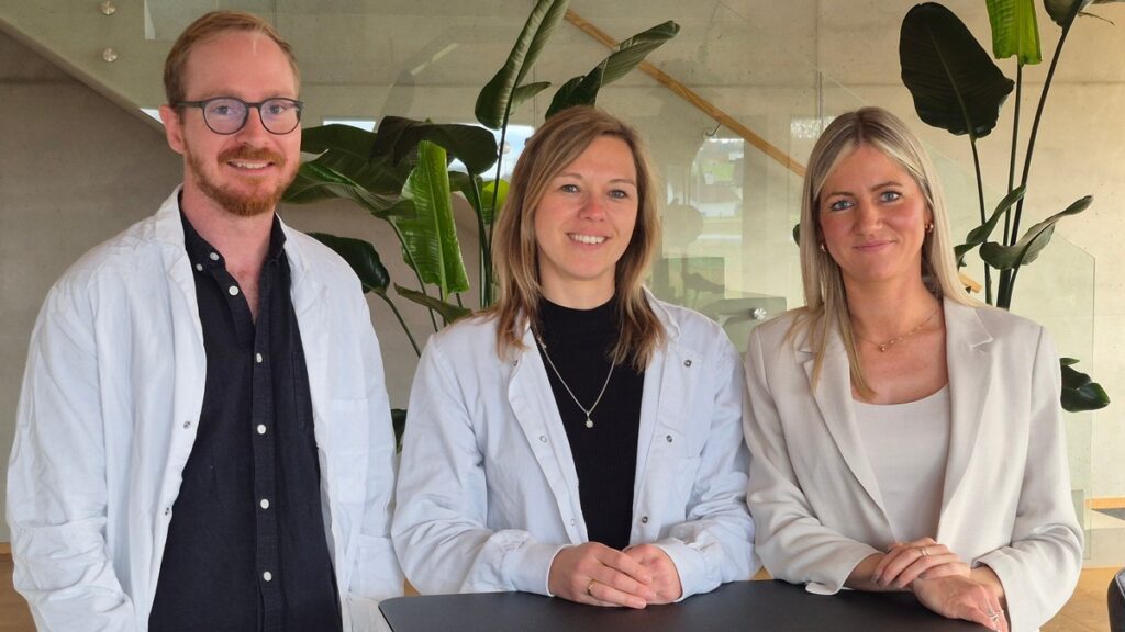 Drei Personen in weißen Kitteln stehen an einem Tisch in einem modernen Innenraum mit Pflanzen im Hintergrund.