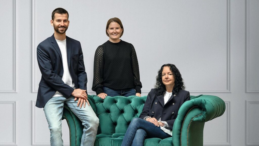 Drei Personen posieren auf einem grünen Chesterfield-Sofa in einem neutralen Raum.