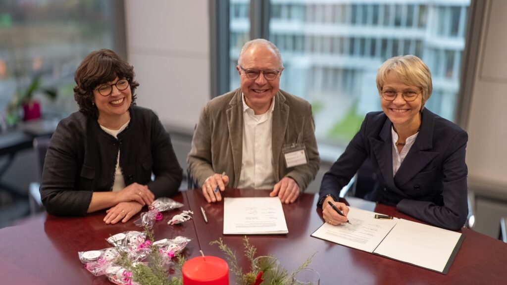 Drei Personen, die an einem Tisch sitzen und Dokumente unterzeichnen, während sie in die Kamera lächeln.