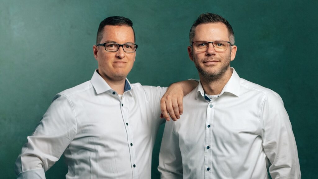 Zwei Männer in weißen Hemden mit Brillen, einer mit Arm auf der Schulter des anderen, vor grünem Hintergrund.