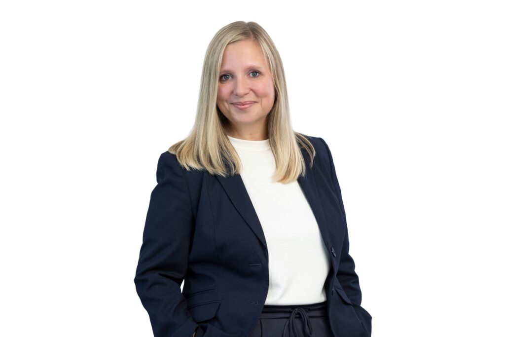 Frau in einem dunkelblauen Blazer und hellem Oberteil mit langen blonden Haaren, die lächelt und in neutraler Pose steht.