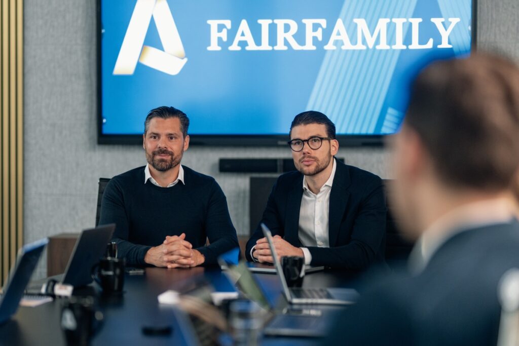 Zwei Männer sitzen an einem Konferenztisch während eines Meetings mit dem Logo von Fairfamily im Hintergrund.