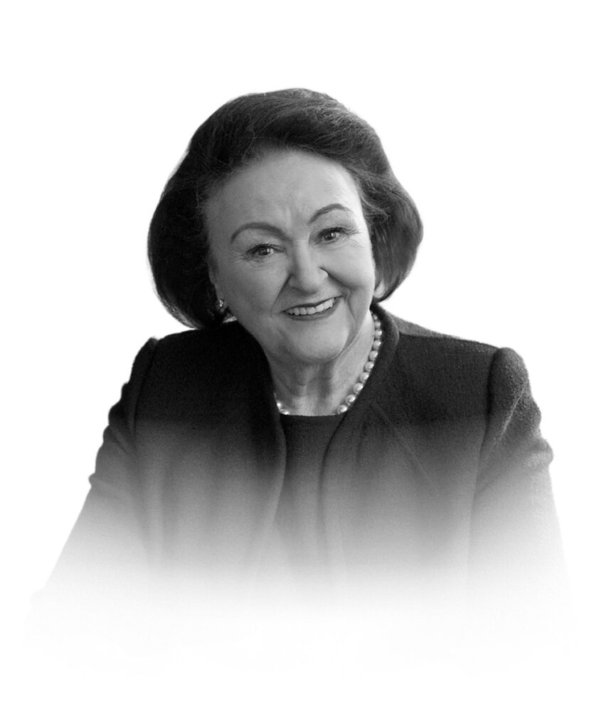 Porträt einer lächelnden Frau mit Perlenkette und dunkler Jacke, in einem neutralen Schwarz-Weiß-Stil.