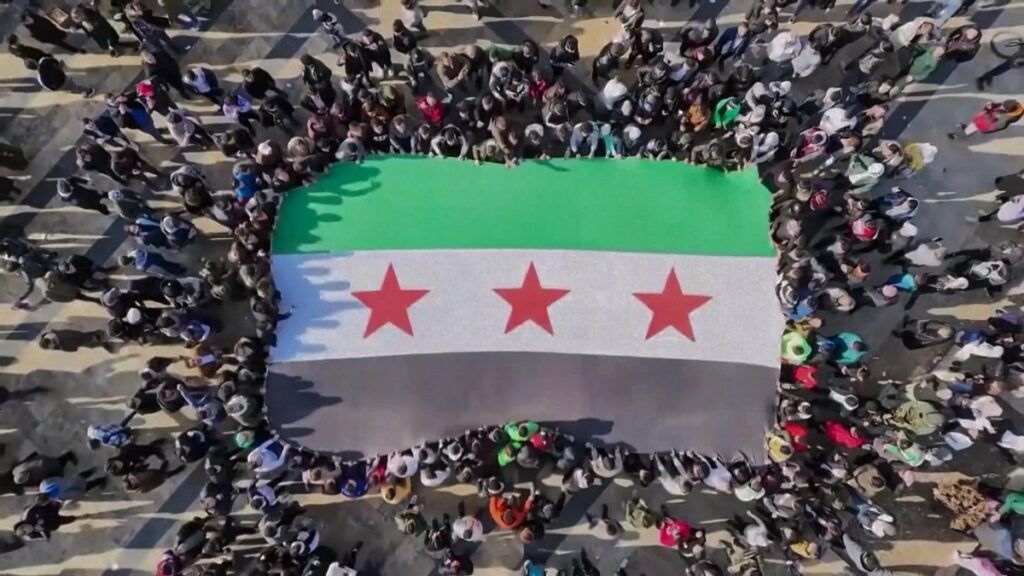 Große Menschenmenge, die eine syrische Flagge mit drei roten Sternen auf dem Boden präsentiert.