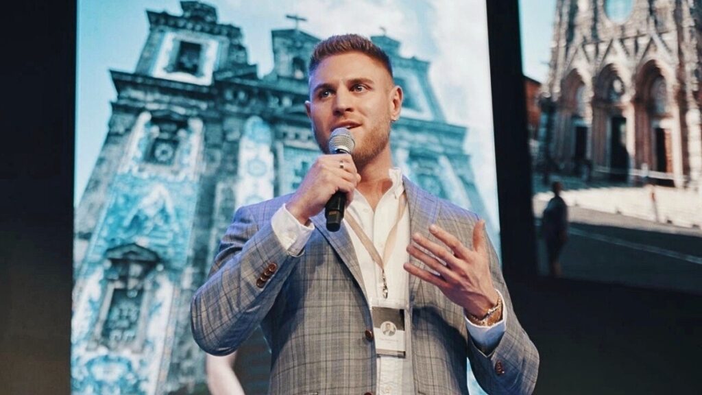 Mann in Anzug spricht mit Mikrofon auf einer Bühne vor einer großen Leinwand mit historischen Gebäuden im Hintergrund.