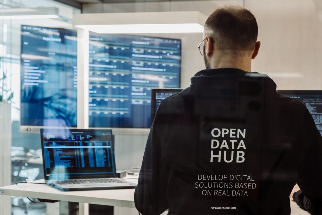 Person in einem Hoodie mit der Aufschrift 'OPEN DATA HUB' vor mehreren Monitoren in einem modernen Büro.