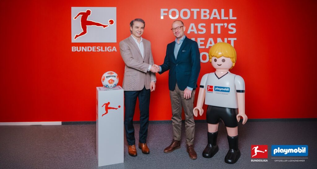 Zwei Männer schütteln sich die Hände vor einer roten Wand mit dem Bundesliga-Logo und einem Playmobil-Figur in Fußballkleidung.