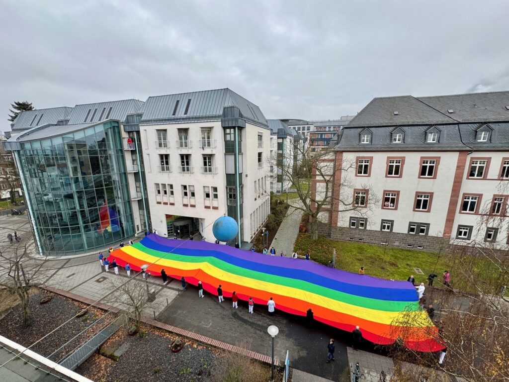 Bunte Regenbogenflagge, die von mehreren Personen vor einem modernen Gebäude gehalten wird.