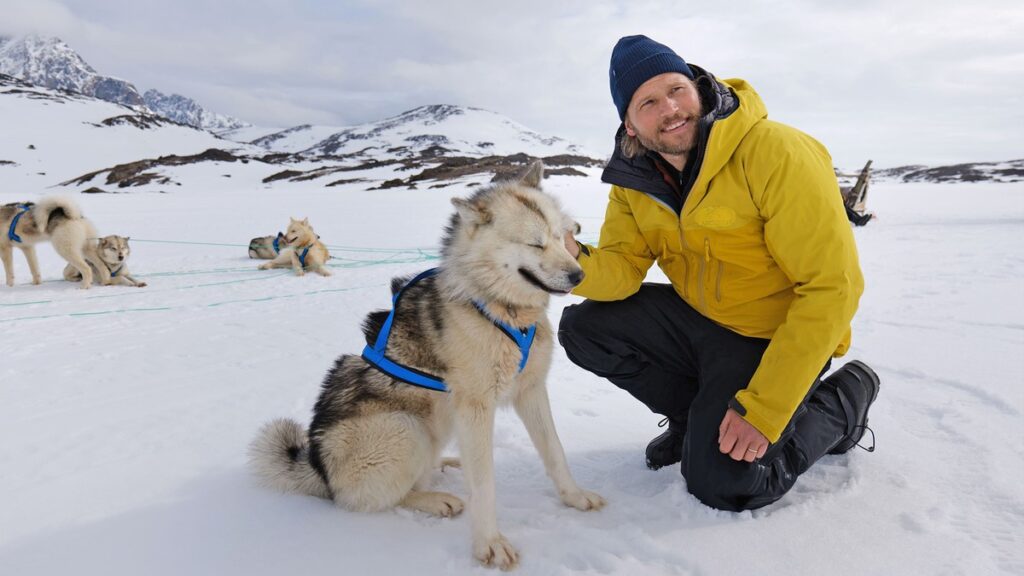 Mann in gelber Jacke kniet neben einem Schlittenhund auf einer Schneefläche in der Arktis.