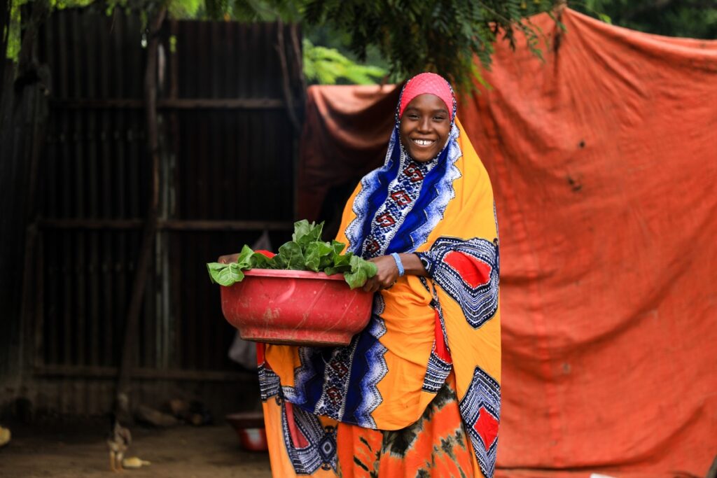 Frau in traditioneller Kleidung hält einen roten Behälter mit frischem Gemüse in der Hand.