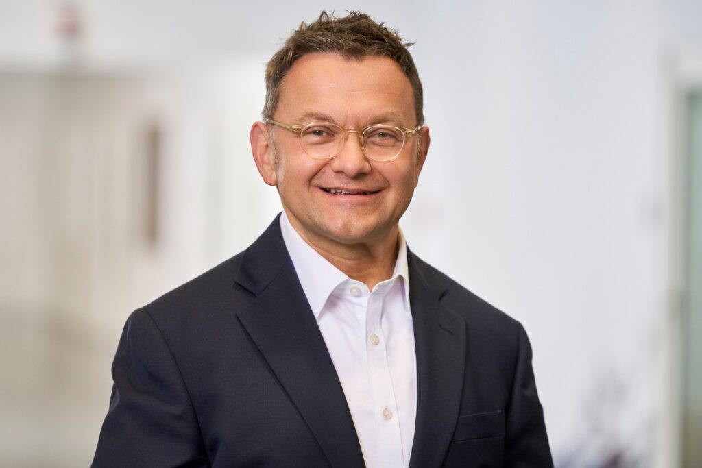 Mann mit Brille und Anzug, der in die Kamera lächelt, vor einem hellen Hintergrund.