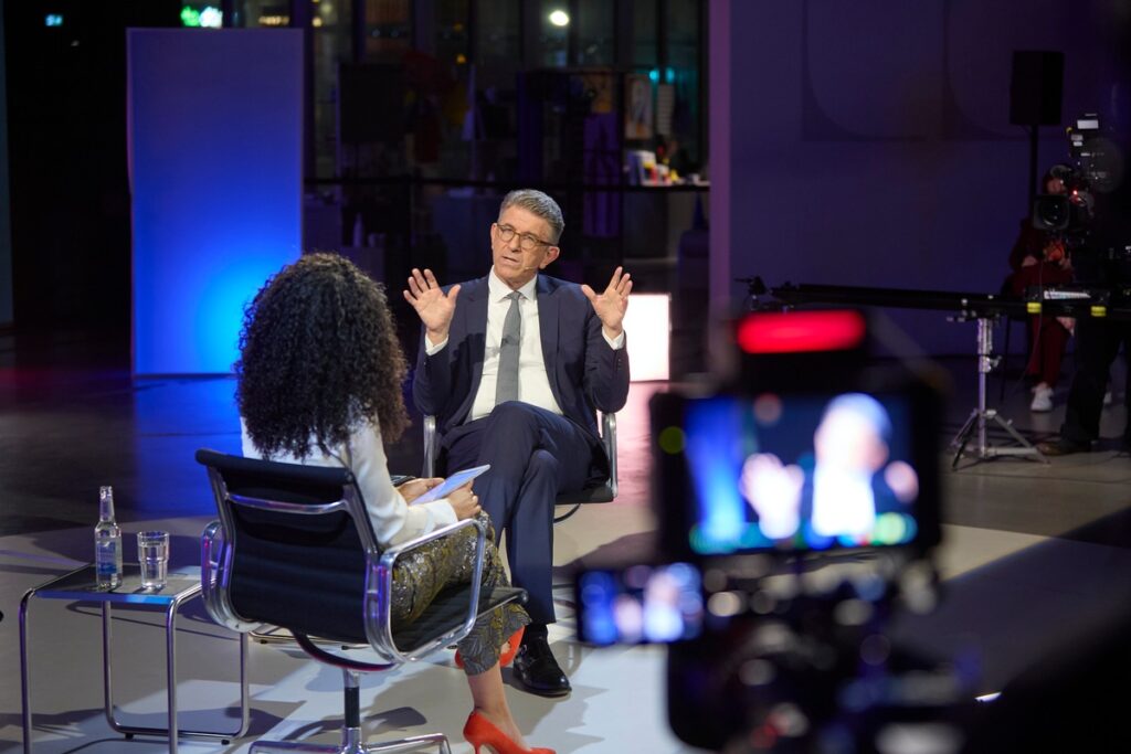 Mann in Anzug sitzt im Interview mit einer Frau, während eine Kamera auf sie gerichtet ist.