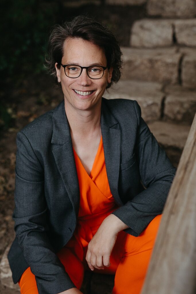 Porträt einer Frau mit Brille und lockigem Haar, die in einem grauen Blazer lächelt.