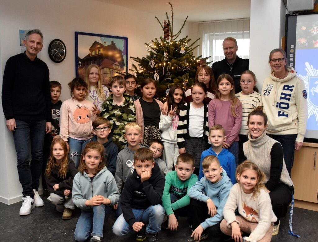 Gruppe von Kindern und Erwachsenen vor einem Weihnachtsbaum in einem Raum, umgeben von Dekorationen und festlicher Atmosphäre.