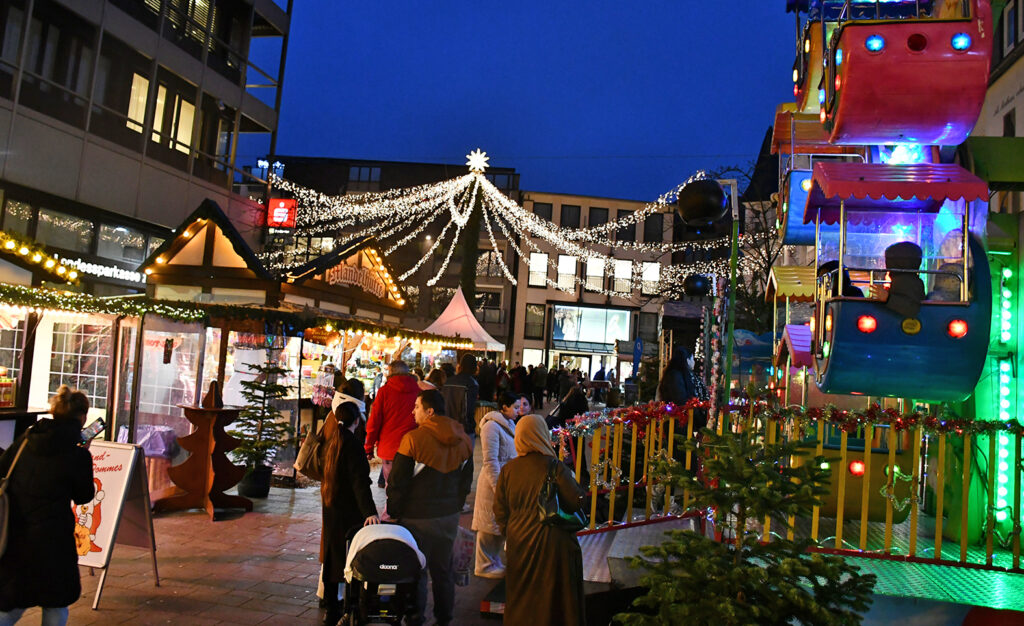 Weihnachtsmarkt mit festlich beleuchteten Ständen und Fahrgeschäften in einer Stadtstraße bei Dämmerung.