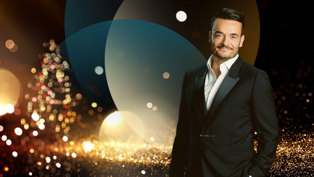 Mann in schwarzem Anzug mit weißem Hemd, lächelnd vor einem festlichen Hintergrund mit Lichtern und einem Weihnachtsbaum.