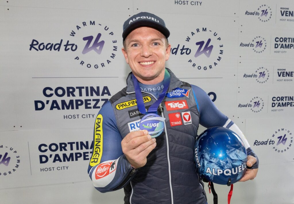 Wolfgang Kindl hält eine Medaille und einen Helm in der Hand, während er vor einem Hintergrund mit dem Logo von Cortina d'Ampezzo steht.