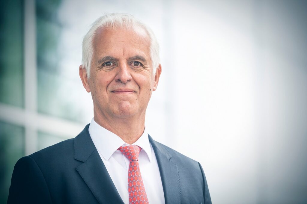 Mann in einem Anzug mit grauen Haaren und roter Krawatte, der in die Kamera lächelt.