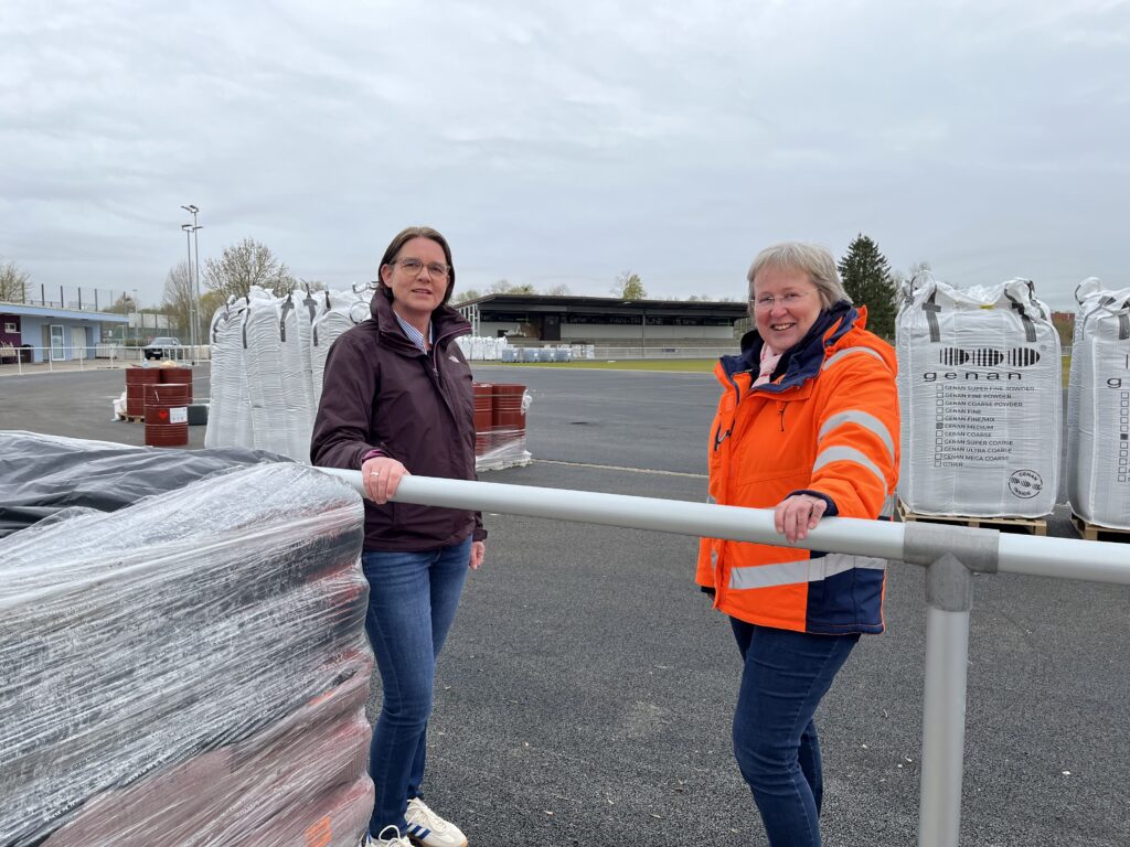 In der Weiling-Arena steht das Material für die Laufbahn-Beschichtung bereit, die Bauarbeiten begleiten (vl.) Karin Hackling (Fachteam Freizeit, Sport) und Renate Rösing (Fachteam FB 70/Tiefbau).