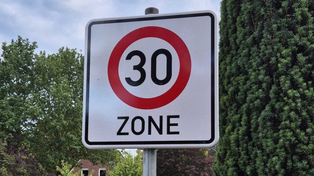 Verkehrsschild Tempo-30-Zone am Straßenrand vor Bäumen und Wohnhäusern