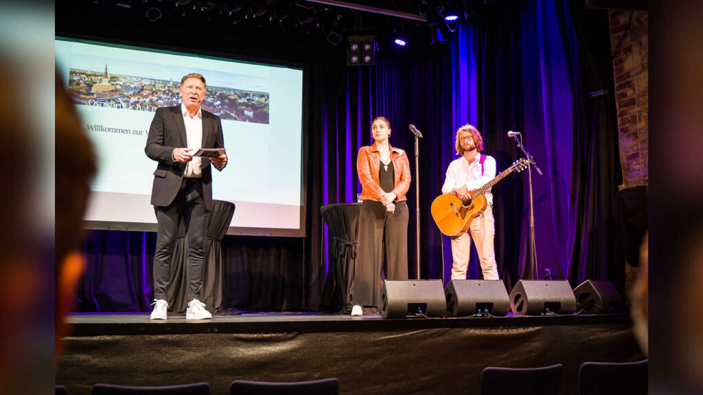 Moderator Ludger Abeln mit einem Musikduo auf der Bühne der Lagerhalle beim Wahlabend 2021