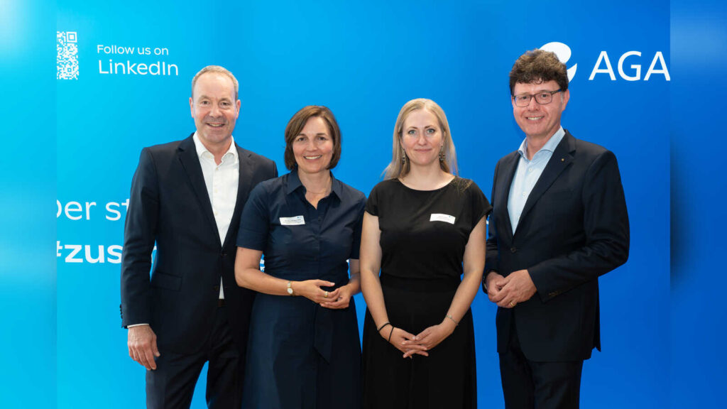 Vier Personen stehen nebeneinander vor einer blauen Wand mit AGA-Logo