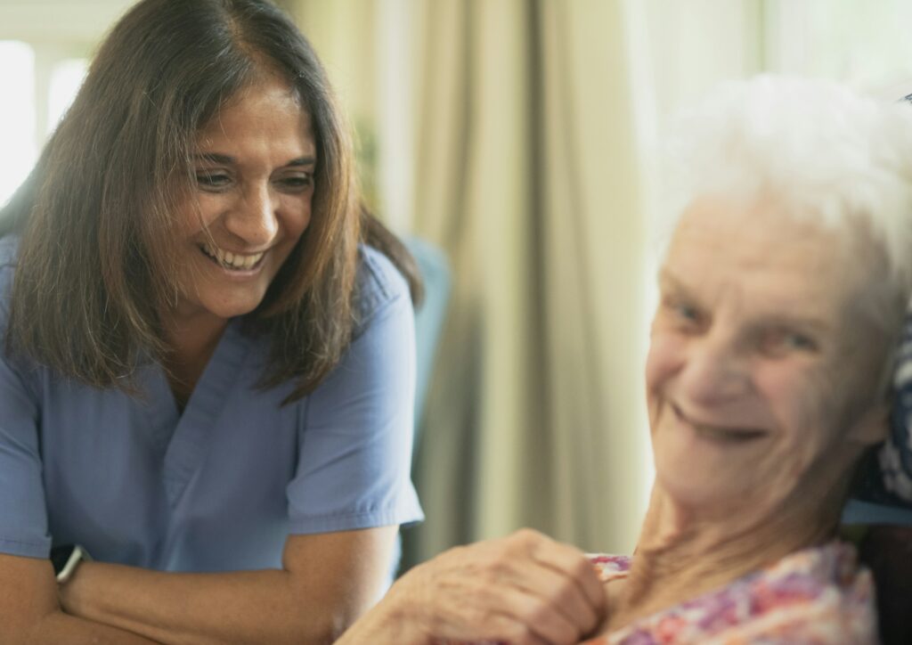 Pflegekraft lächelt mit älterer Patientin im Zimmer, Symbolbild zum Pflege-Modellprojekt in Emsland und Grafschaft Bentheim