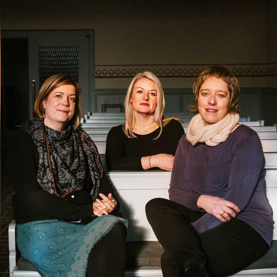 Drei Frauen sitzen in Kirchenbänken und blicken in die Kamera