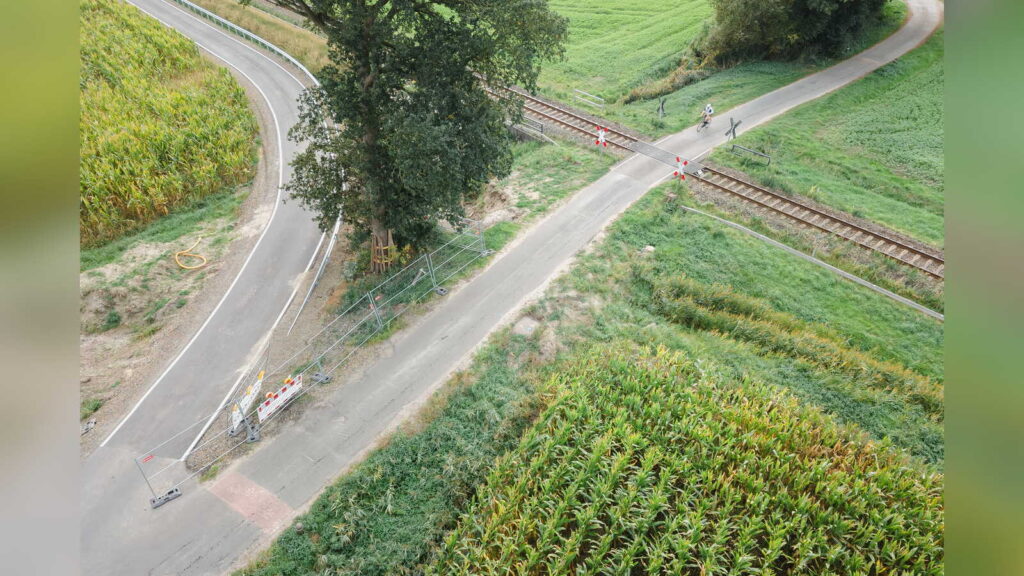 Luftbild des unbeschrankten Bahnübergangs Brettberger Aue in Lohne-Zerhusen mit Andreaskreuzen, einem Radfahrer auf dem Übergang, Absperrgittern und der neu asphaltierten Straße, die links im Bogen abzweigt